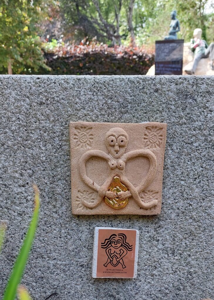 Ceramic Sheela na Gig plaque with gold vulva on a stone bench in Merrion Square, Her son Oscar's statue can be glimpsed in the background. 