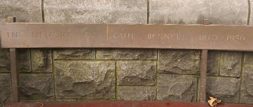 Seat with metal back against a stone wall in St Stephen's Green, Dublin. Engraved with text 'In Memory of Louie Bennett'.