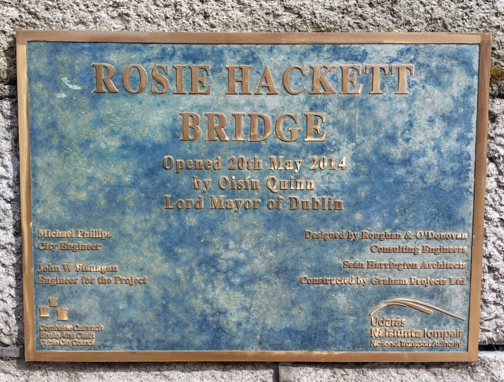 Rectangular metal plaque against a stone wall. Text in bronze reads Rosie Hackett bridge opened 20th May 2014 by Oisín Quinn, Lord Mayor of Dublin and a list of engineers, architects and construction firms involved in the project.