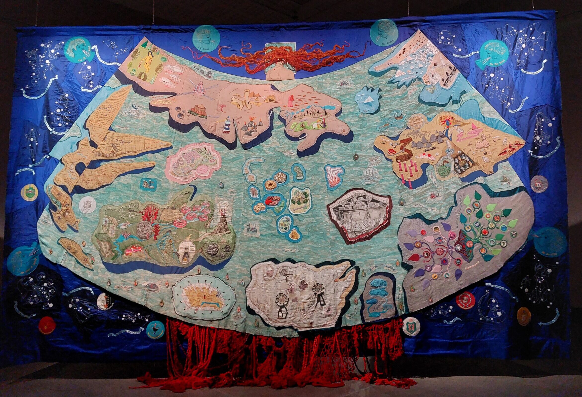 Photo of The Map, an art piece featuring hand embroidered images on a large quilt by Rachel Fallon and Alice Maher. The photo shows the whole map so the detail isn't clear but it appears as a blue sky background, with land masses on a sea of green.