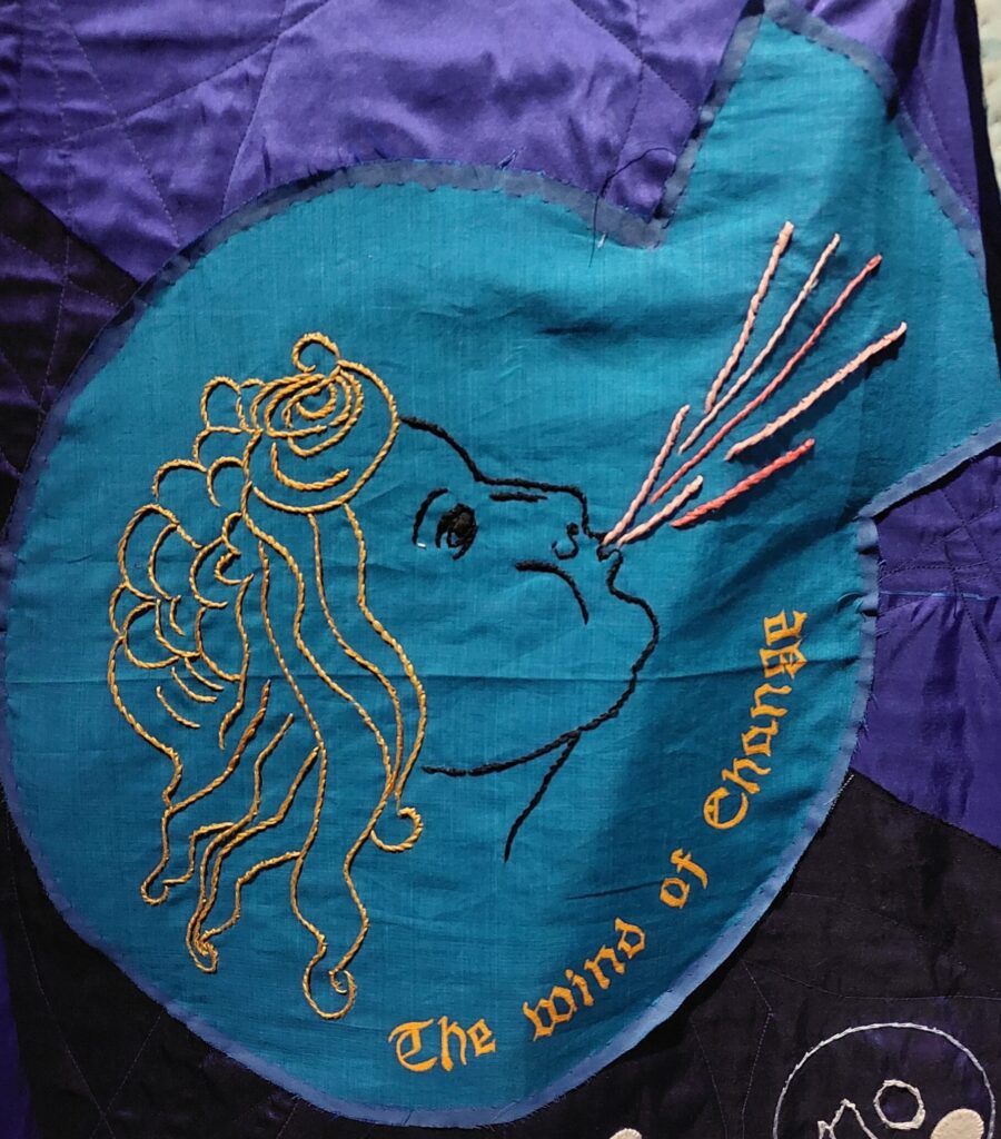 Embroidered panel of a female face blowing, with the words the wInd of change