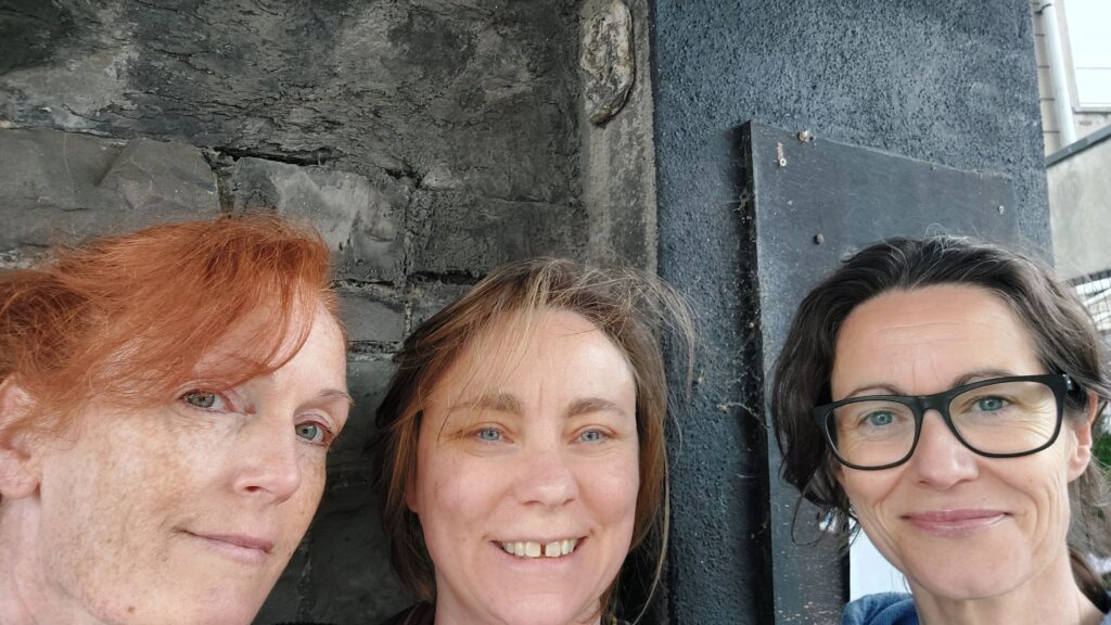 three white women stand below a very small Sheela na Gig on a wall in Grangegorman, Dublin