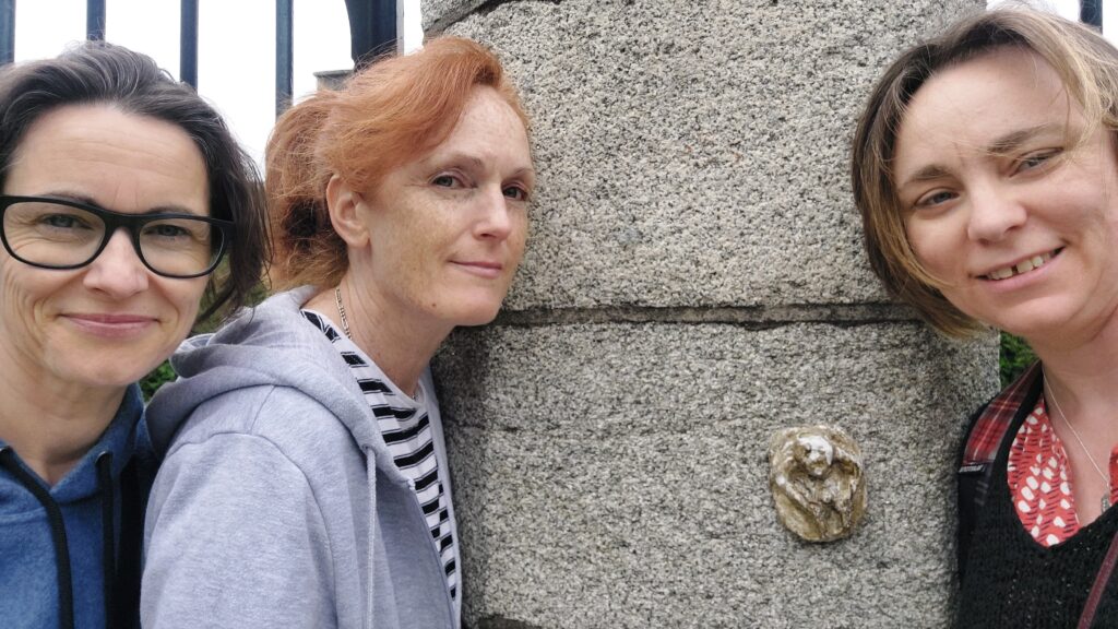 Three white women stand around a small Sheela na Gig on the gates of Arbour Hill prison, Dublin