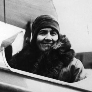 Black and white photo of aviator Sophie Peirce, a white woman sitting in the cockpit of a c1920s plane, wearing a cloche hat, a fur wrap and a smile.