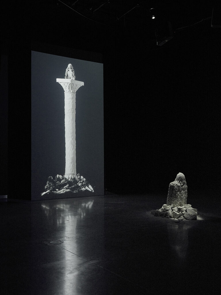 Scene from The Tower by Jesse Jones. Black and white still shows a darkened room with an illuminated statue of a woman kneeling, facing an screen with and image of a cross.