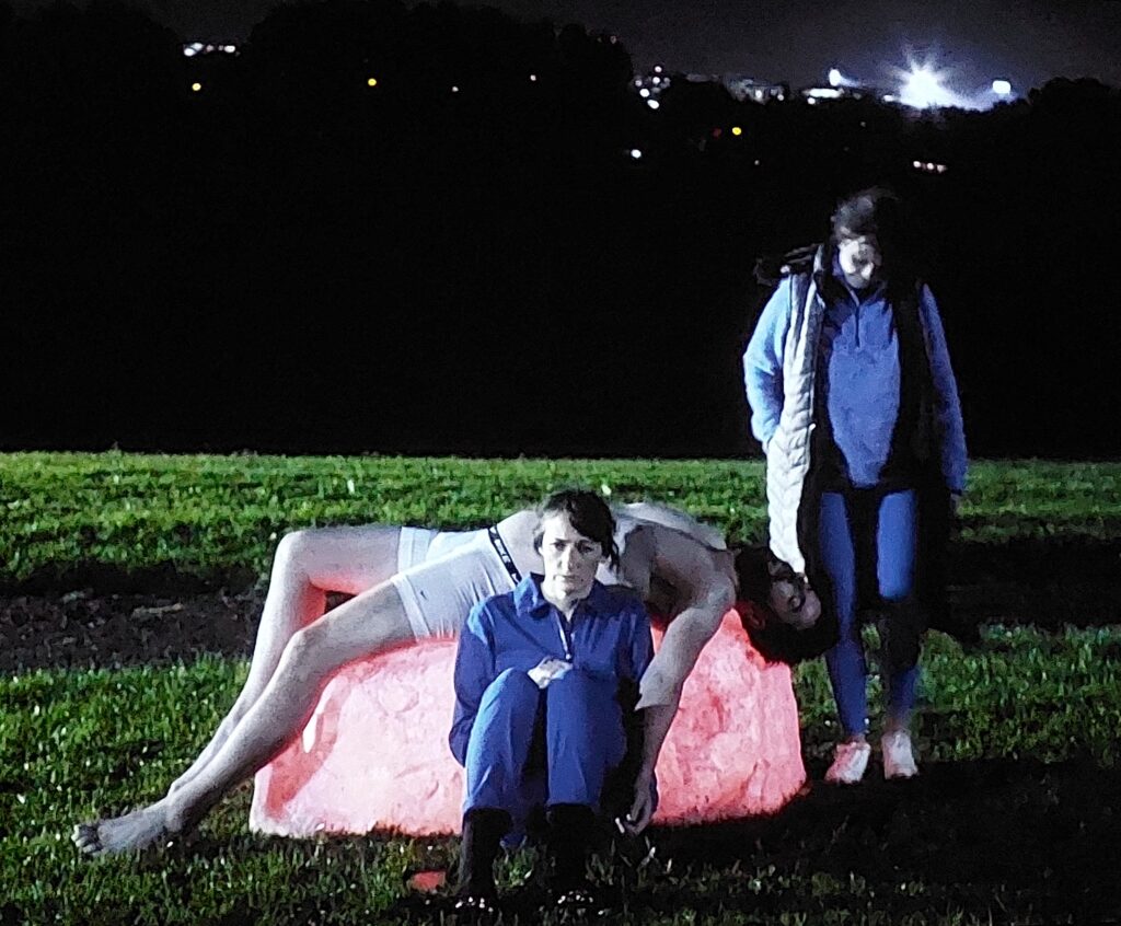 Still from A Mary Magdalene Experience by Grace Dyas. A man lies draped across a glowing rock of pink quartz while his mother sits forlornly in front of him and another woman stands to the side, suggesting a Pieta 