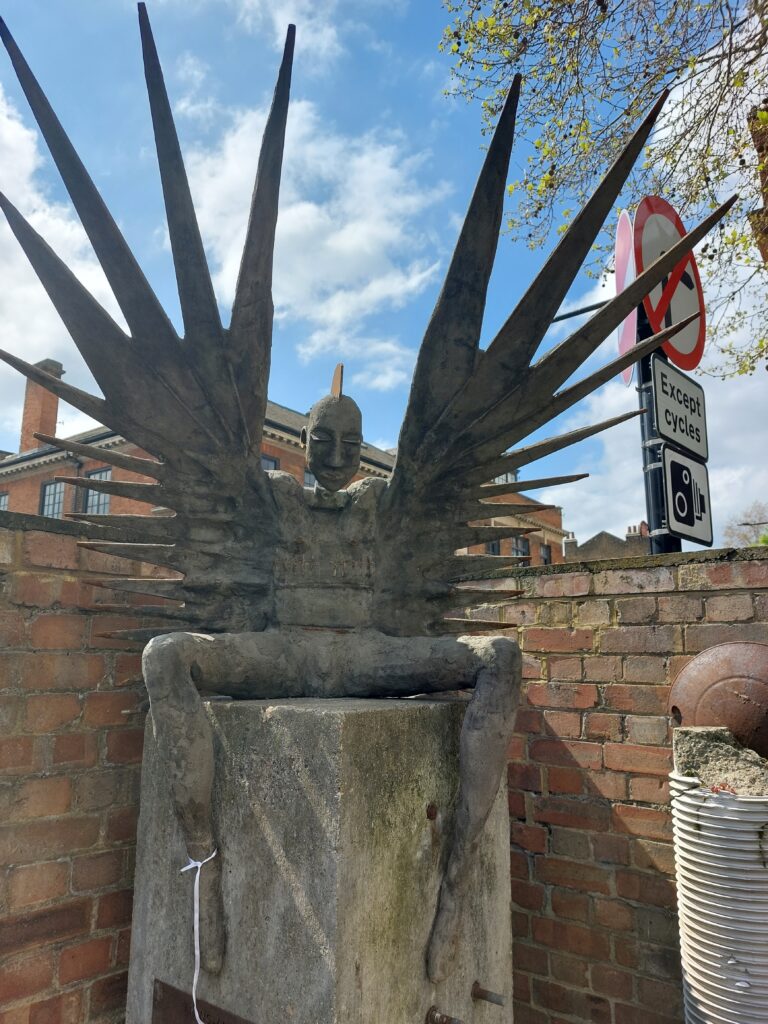 A steel and concrete sculpture of a trans angel, with both male and female parts and jagged wings, in Crossbones Graveyard, Southwark