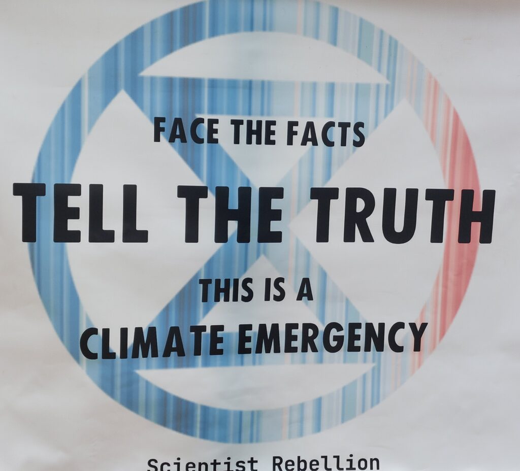 Poster at Extinction Rebellion event with text on the background of an hourglass: face the facts, tell the truth, this is a climate emergency.