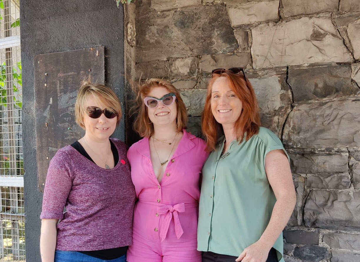 Two of the Wild Gees with comedian Blathin de Burca (three smiley white women) standing in front of a tiny Sheela na Gig on a wall in Grangegorman, Dublin