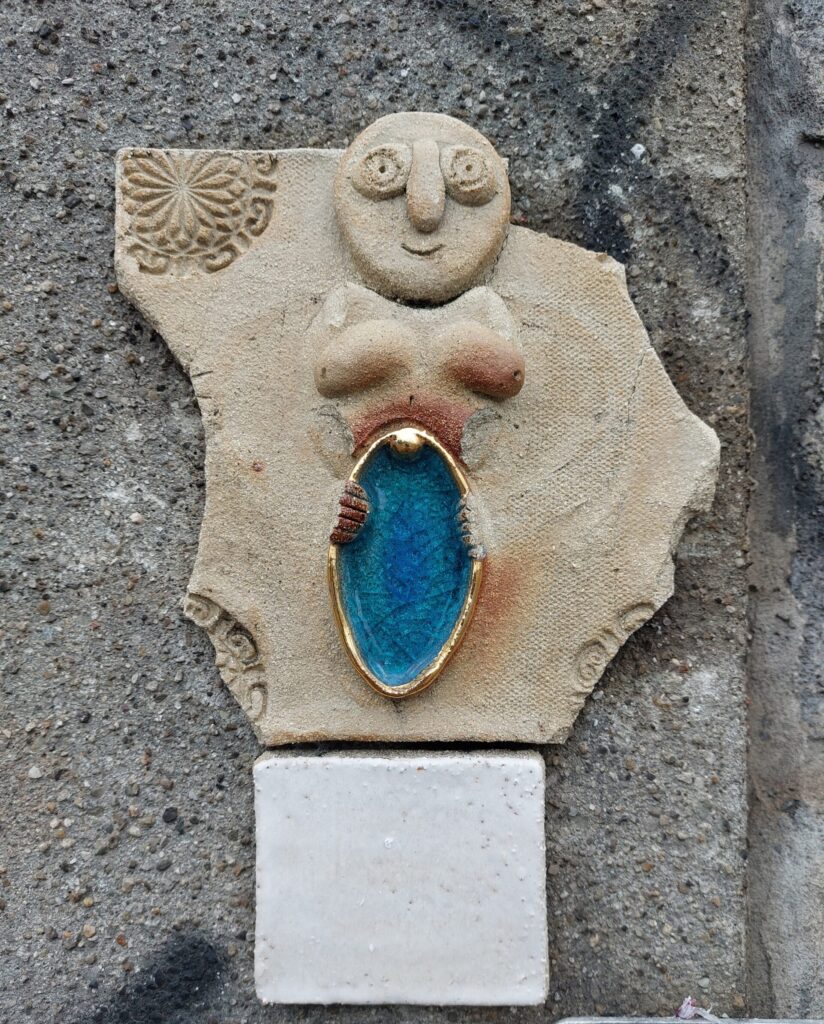 Ceramic Sheela na Gig figure with blue glass vulva. The figure is missing arms and the corners of the tile it sits on, which is attached to a stone wall.