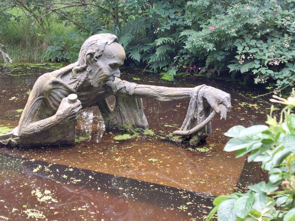 Sculpture in water of a gaunt figure reaching for something. The pond is in a woodland and is surrounded by greenery